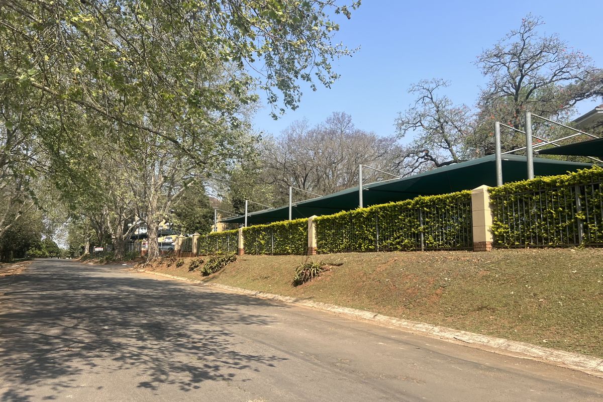 Street view of the office property perimeter, featuring secure metal fencing, brick pillars, and overhead shade netting structures along the roadside.