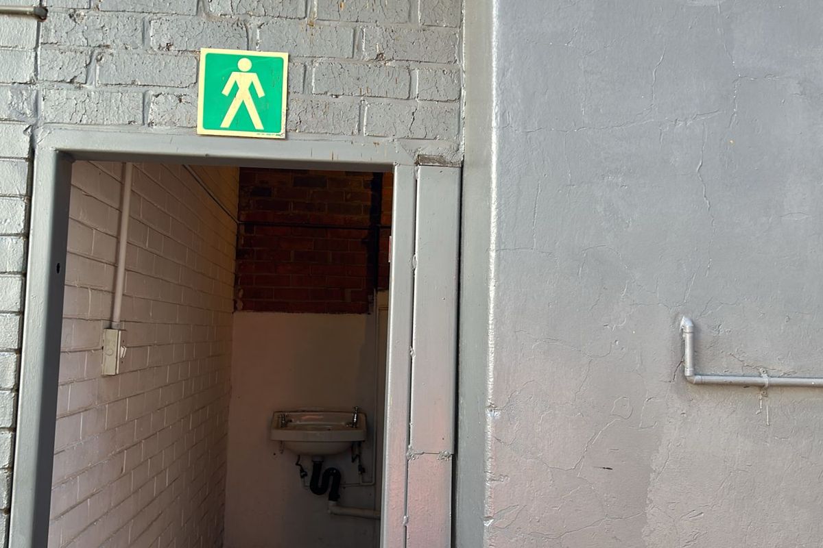 A utilitarian washroom area featuring a wall-mounted basin, exposed brickwork, and industrial-style painted masonry walls with an emergency exit sign.