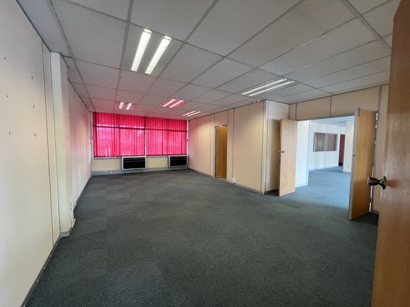 An empty, open-plan office space featuring a suspended ceiling, recessed lighting, carpeted flooring, and built-in air conditioning units with red window blinds.