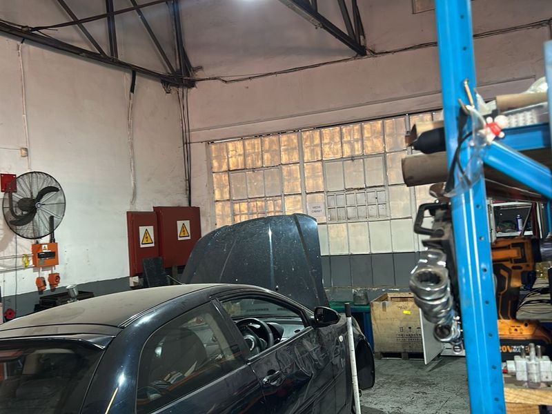 Commercial workshop space featuring high ceilings, industrial lighting, wall-mounted ventilation fan, and large multi-pane windows providing natural light.