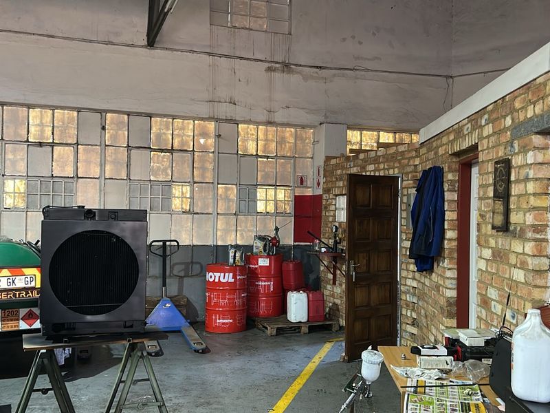 Industrial workshop space featuring high ceilings, large multi-pane windows, exposed brick walls, and concrete flooring suitable for commercial or light industrial operations.
