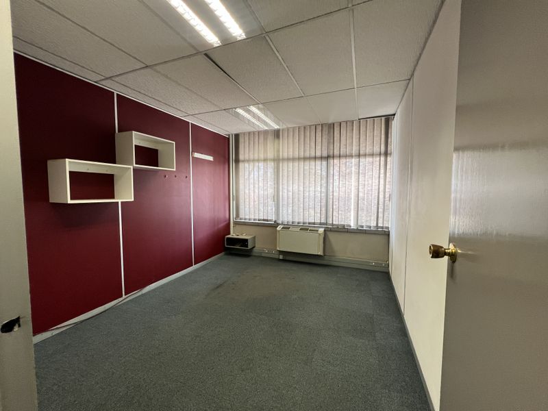 A vacant office space featuring carpeted flooring, a suspended ceiling with integrated lighting, vertical blinds, and a wall-mounted HVAC unit.