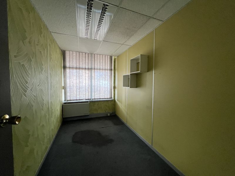 A small, vacant office space featuring a suspended ceiling, HVAC unit, and vertical blinds, currently requiring cosmetic updates and flooring replacement.
