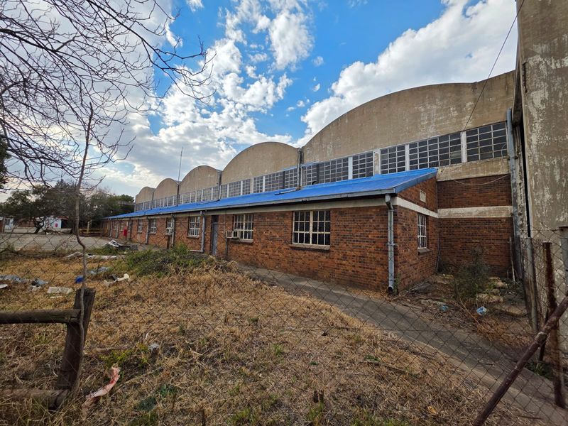 Exterior view of a large industrial building featuring a distinctive barrel-vaulted roof, brick facade, and multi-pane windows, set within a fenced property.