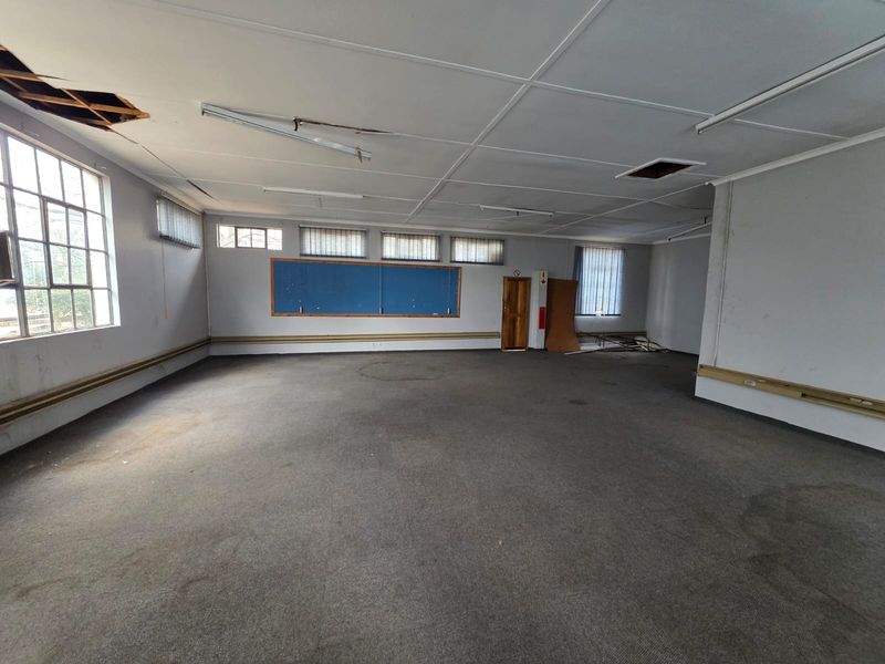 Spacious open-plan office or classroom area featuring large windows, wall-mounted display board, and carpeted flooring, requiring significant maintenance and refurbishment.