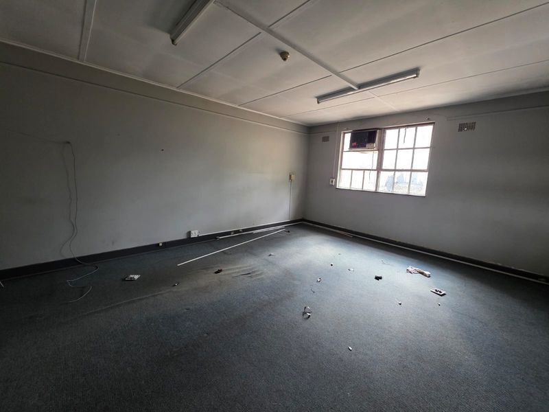 An empty office space featuring carpet flooring, a window-mounted air conditioning unit, and fluorescent lighting fixtures.