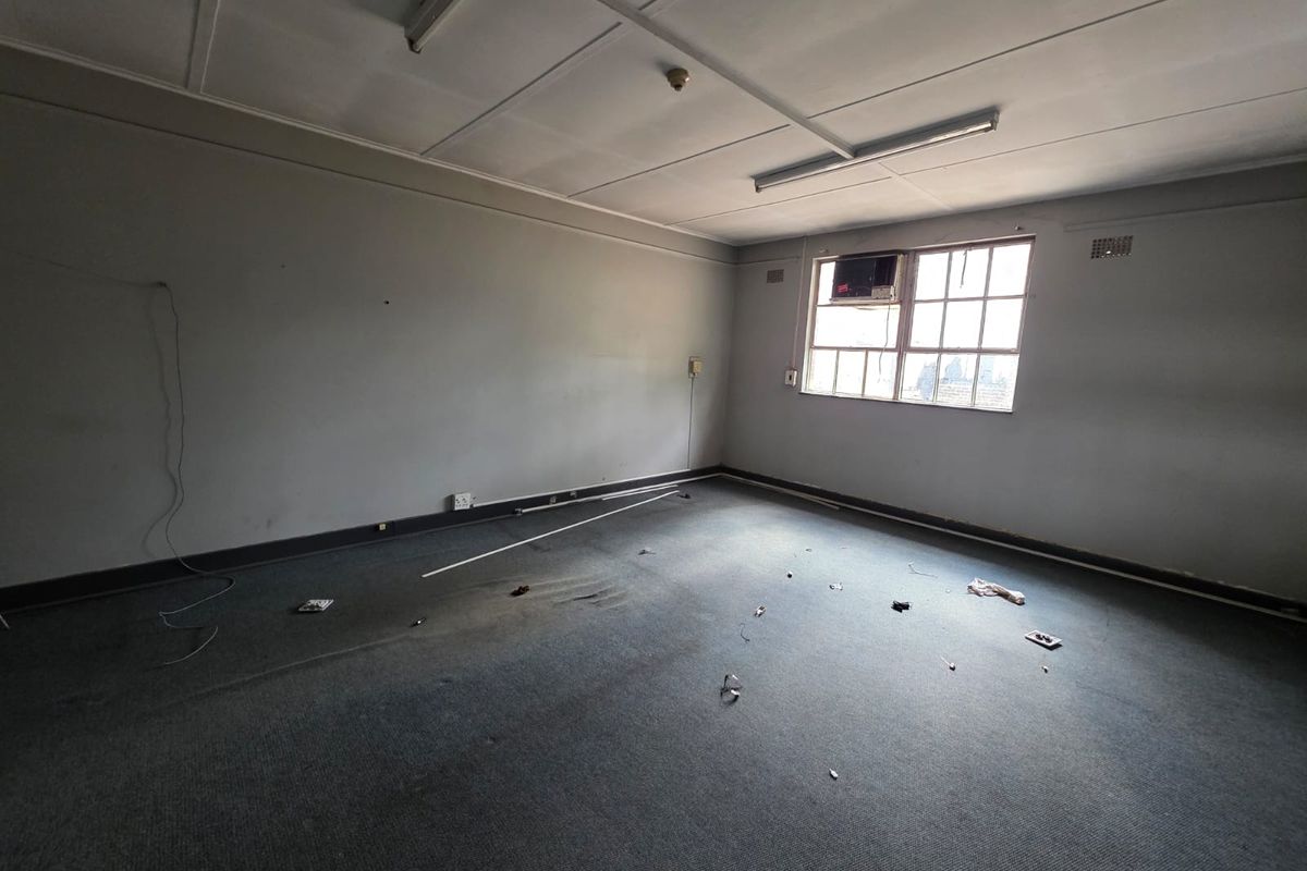 An empty office space featuring carpet flooring, a window-mounted air conditioning unit, and fluorescent lighting fixtures.