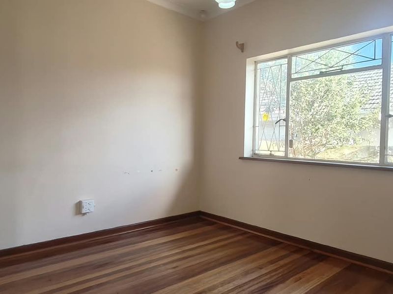A bright, empty office space featuring polished wooden flooring, neutral walls, and a large window with security bars.