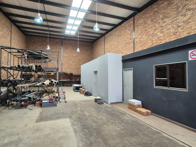 Spacious industrial utility area featuring exposed brick walls, high ceilings with steel trusses, skylights, and a dedicated internal office partition.