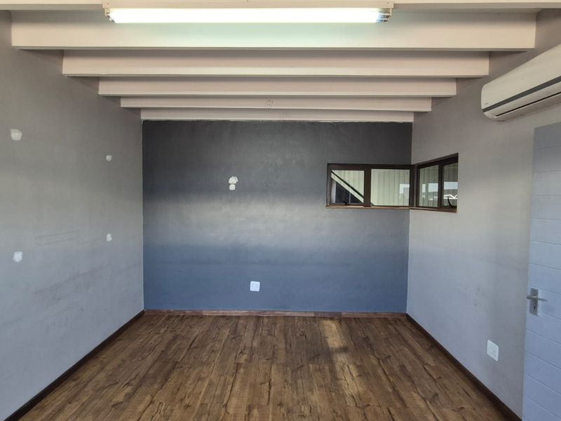 A clean, empty office space featuring exposed ceiling beams, laminate flooring, a wall-mounted air conditioning unit, and corner windows for natural light.
