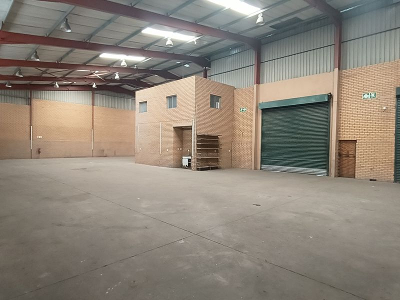 Spacious industrial warehouse featuring high ceilings with exposed steel trusses, concrete flooring, multiple roller shutter doors, and an internal mezzanine office structure.