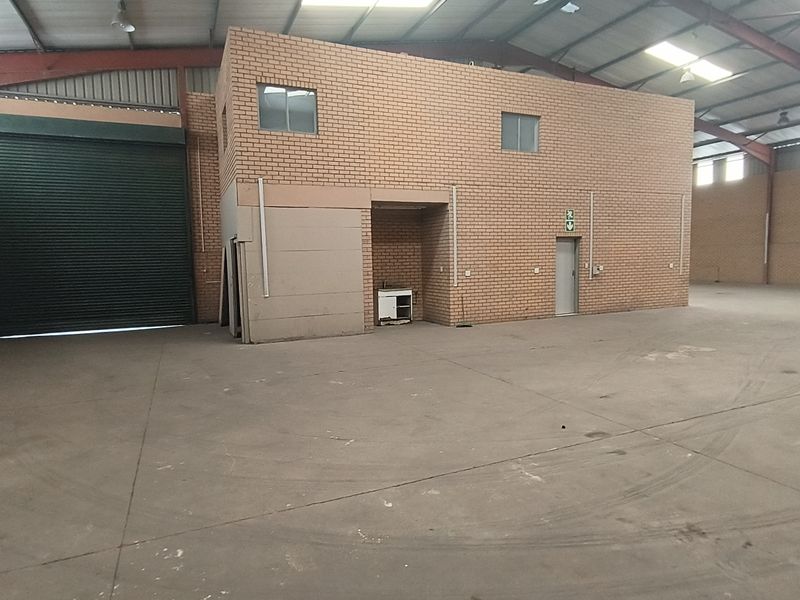 Spacious industrial warehouse unit featuring high ceilings with exposed steel trusses, concrete flooring, a large roller shutter door, and an internal brick office structure.