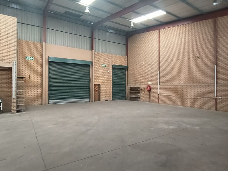 Spacious industrial warehouse unit featuring high ceilings with exposed steel trusses, concrete flooring, face brick walls, and multiple roller shutter doors for easy access.
