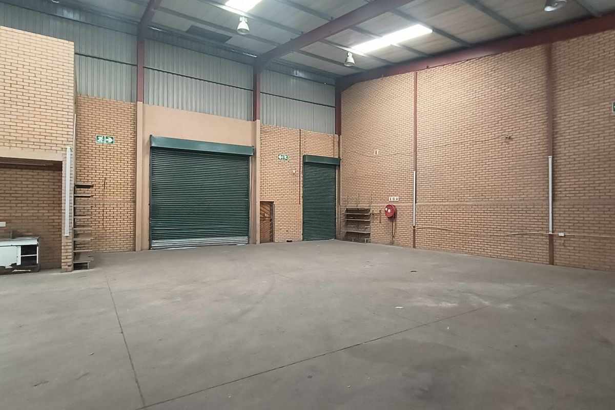 Spacious industrial warehouse unit featuring high ceilings with exposed steel trusses, concrete flooring, face brick walls, and multiple roller shutter doors for easy access.