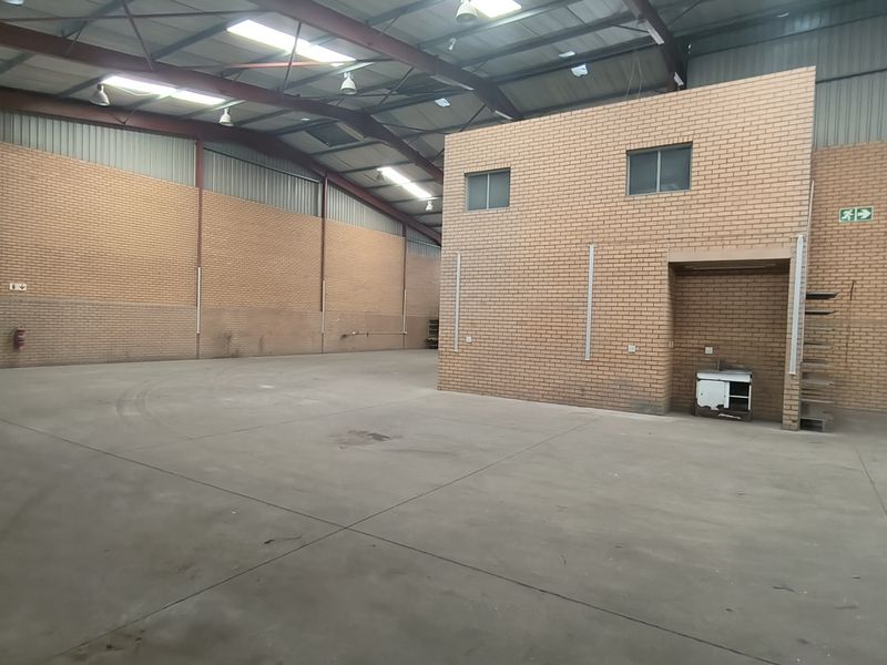 Spacious industrial warehouse featuring high ceilings with exposed steel trusses, concrete flooring, brick walls, and a built-in mezzanine office structure.