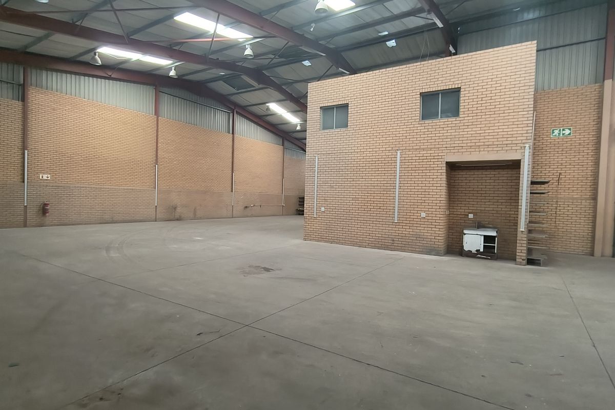 Spacious industrial warehouse featuring high ceilings with exposed steel trusses, concrete flooring, brick walls, and a built-in mezzanine office structure.