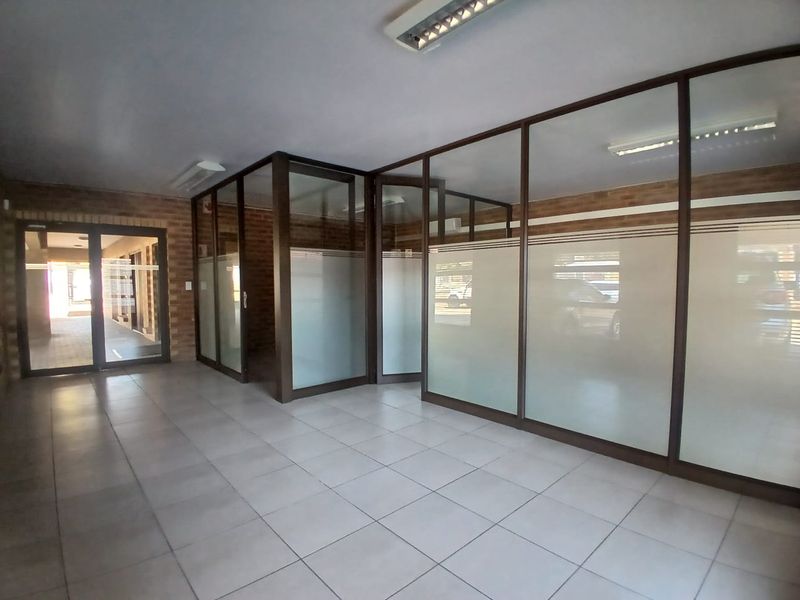 A vacant commercial office space featuring glass partition walls, ceramic tiled flooring, and exposed brick accents, suitable for a professional workspace.