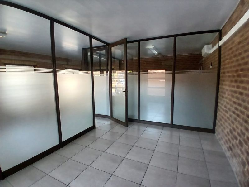 A vacant commercial office space featuring glass partition walls, tiled flooring, an exposed brick wall, and an integrated air conditioning unit.