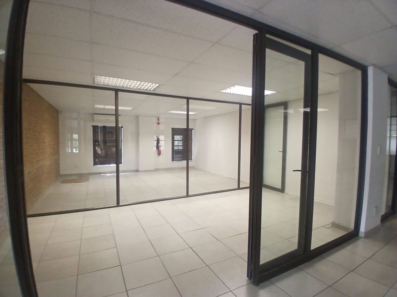A bright, vacant office space featuring modern glass partitions, tiled flooring, and a suspended ceiling with recessed lighting.