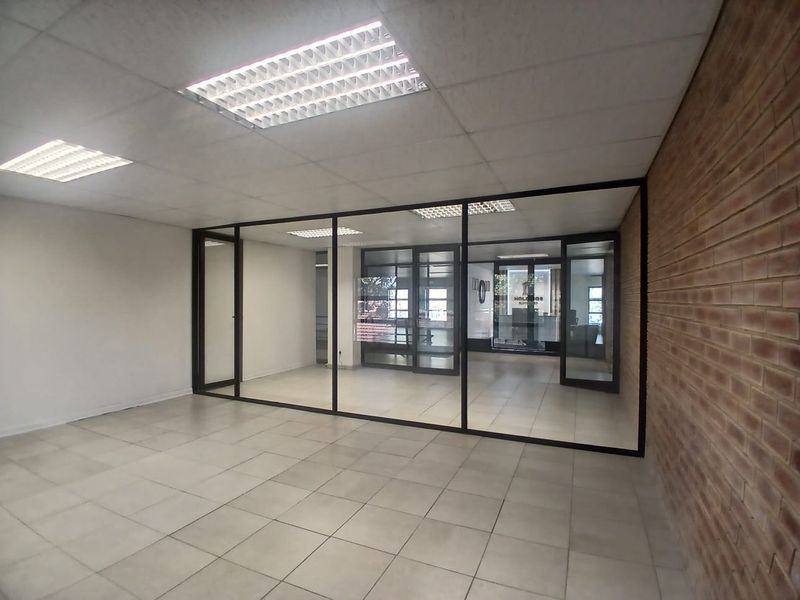 A bright, open-plan office space featuring modern glass partitions, tiled flooring, a feature brick wall, and recessed lighting in a suspended ceiling.