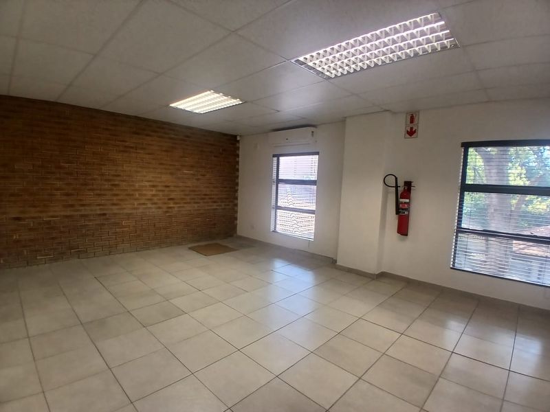 A vacant office space featuring an exposed brick feature wall, tiled flooring, suspended ceiling with recessed lighting, and a wall-mounted air conditioning unit.