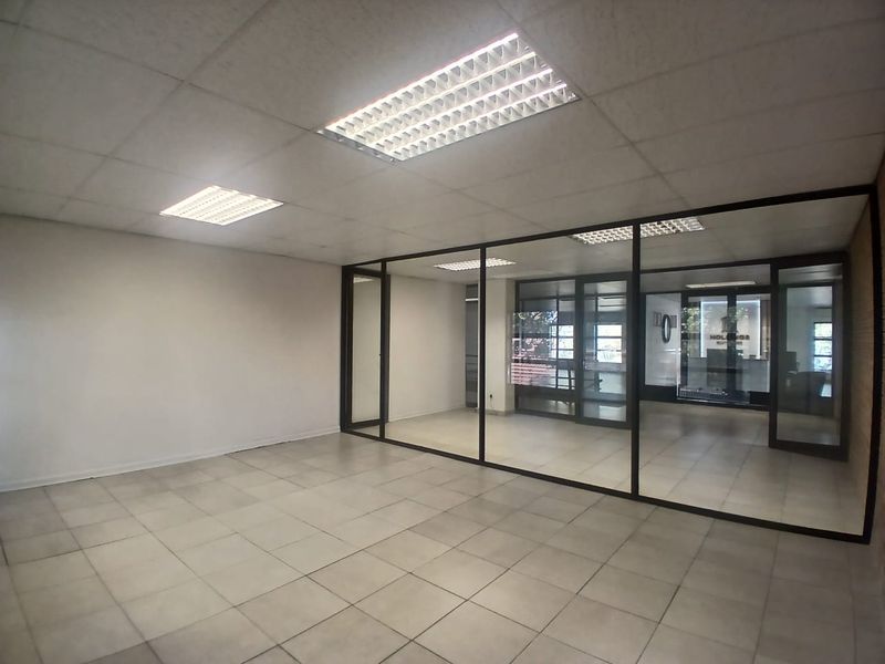 A bright, empty office space featuring modern glass partitions, tiled flooring, and a suspended ceiling with recessed lighting, offering a clean professional environment.