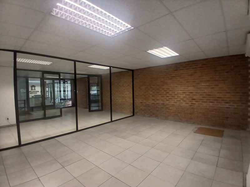 An empty office space featuring a prominent exposed brick wall, glass partitioning, suspended ceiling with recessed lighting, and tiled flooring.