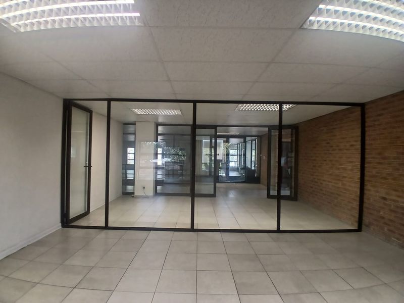 An empty office space featuring tiled flooring, a suspended ceiling with recessed lighting, and a glass partition wall with an exposed brick feature wall.