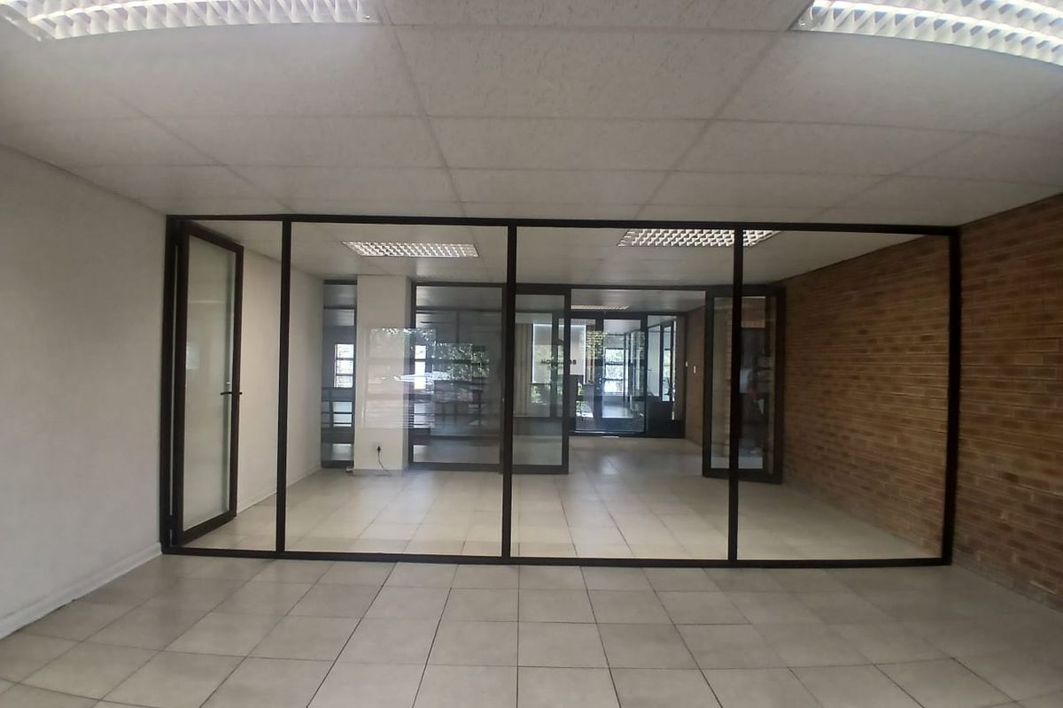 An empty office space featuring tiled flooring, a suspended ceiling with recessed lighting, and a glass partition wall with an exposed brick feature wall.
