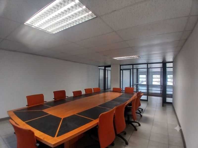 A professional boardroom featuring a large conference table, suspended ceiling with recessed lighting, and glass partition walls providing an open office feel.