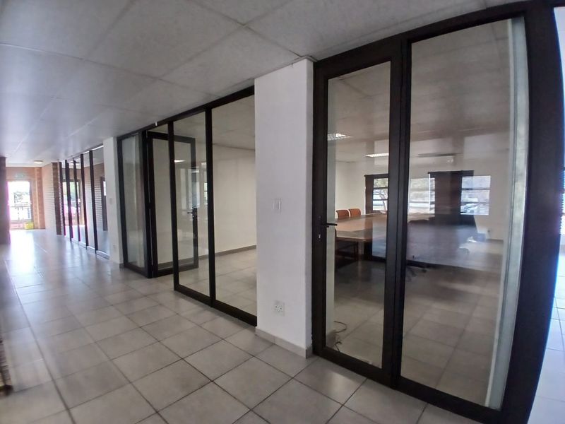 A bright office hallway featuring glass partition walls, tiled flooring, and a suspended ceiling, providing access to multiple private office spaces.