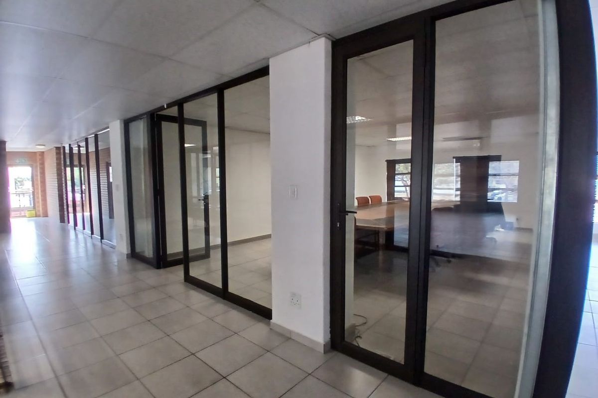 A bright office hallway featuring glass partition walls, tiled flooring, and a suspended ceiling, providing access to multiple private office spaces.