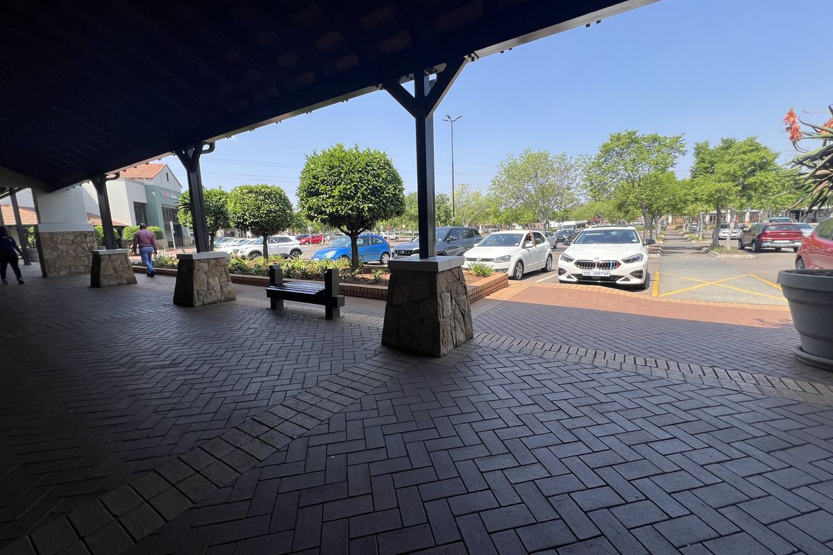 A covered outdoor walkway featuring herringbone brick paving and stone-clad pillars, overlooking a commercial parking area with landscaped garden beds.