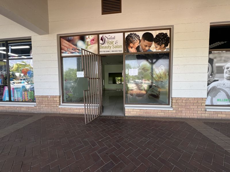 Front entrance of a commercial salon unit featuring a glass storefront, security gate, and brick facade detailing in a retail center.