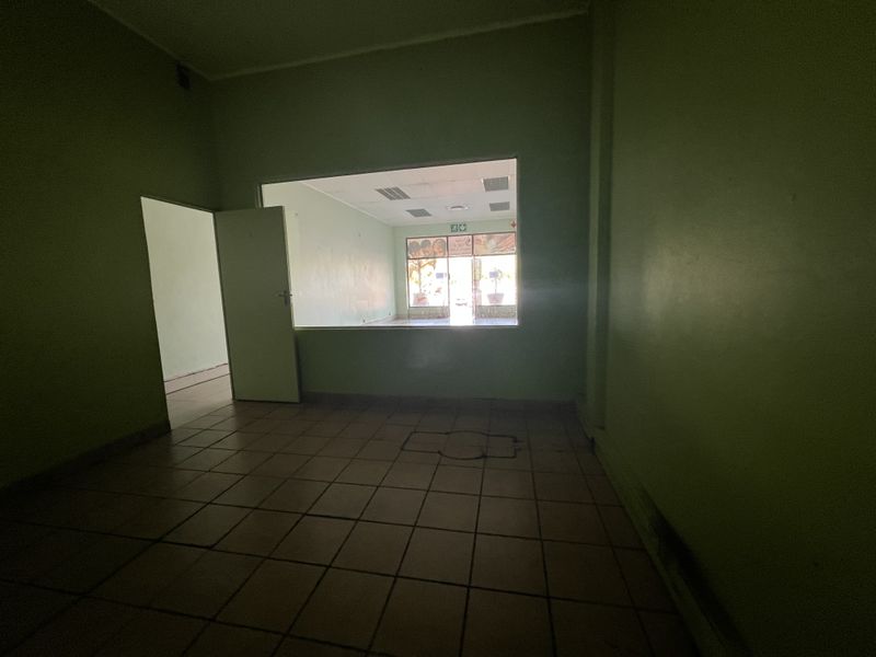 An empty, open-plan commercial space featuring tiled flooring, a drop ceiling, and an internal service window looking through to the front entrance area.