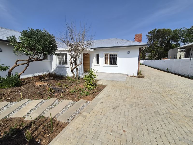 Front exterior view of a well-maintained property featuring a paved driveway, white facade, metal roofing, and a landscaped garden area.