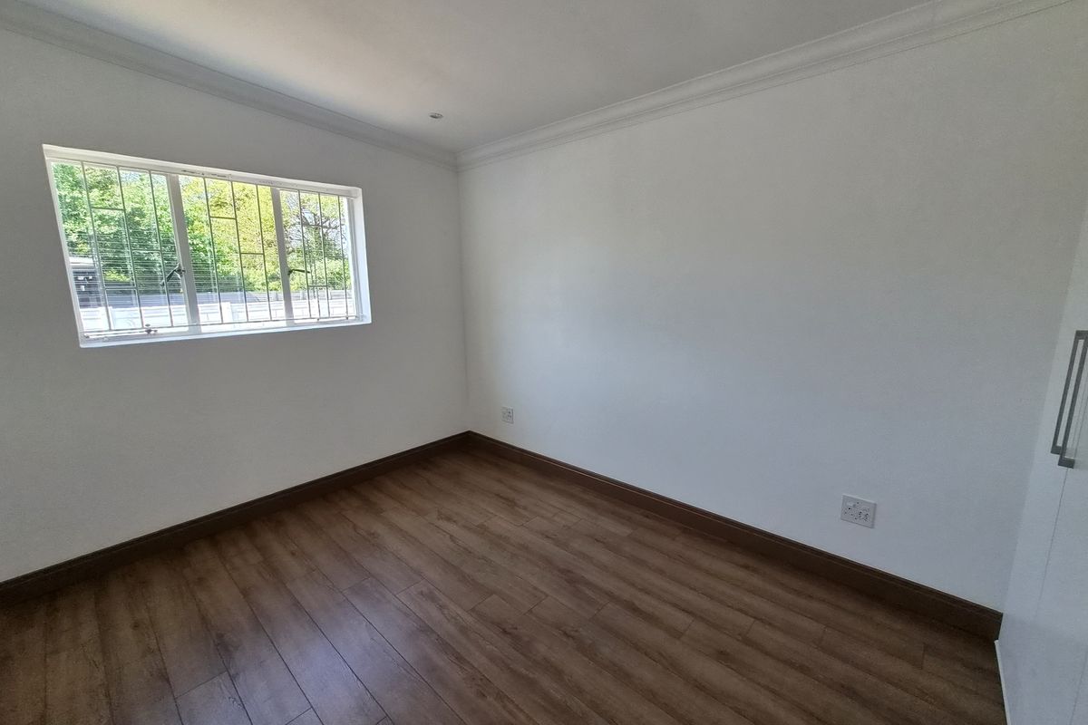 A bright, empty bedroom featuring laminate flooring, built-in cupboards, and a window fitted with security bars.