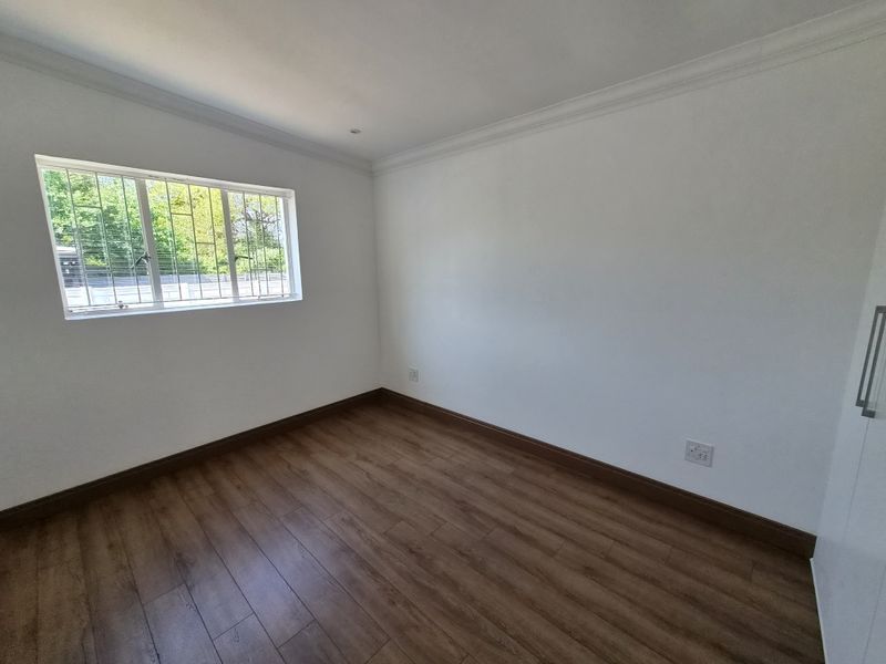 A bright, empty bedroom featuring laminate flooring, built-in cupboards, and a window fitted with security bars.