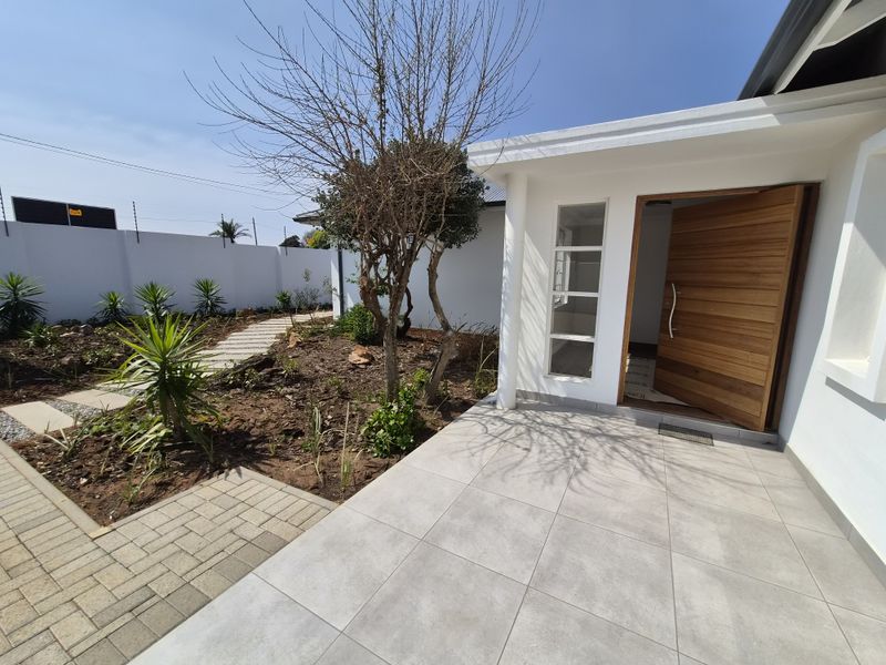 Modern front entrance featuring a large timber door, tiled patio area, and a landscaped garden with paved walkways and secure perimeter walling.