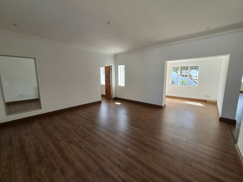 A spacious, empty living area featuring modern laminate flooring, recessed downlighting, and a built-in wall mirror, leading into an adjacent sunlit space.