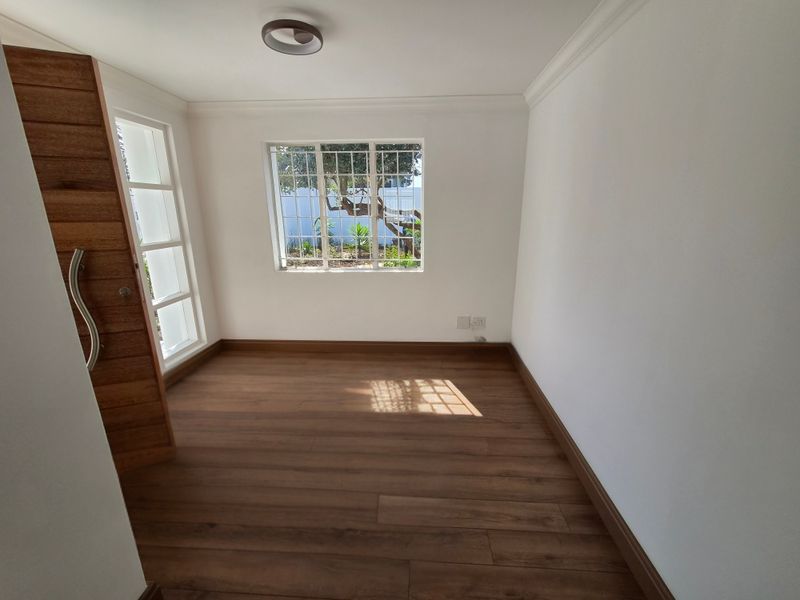 A bright, empty office space featuring modern laminate flooring, a timber door, and a window with security bars, offering a clean and neutral workspace.