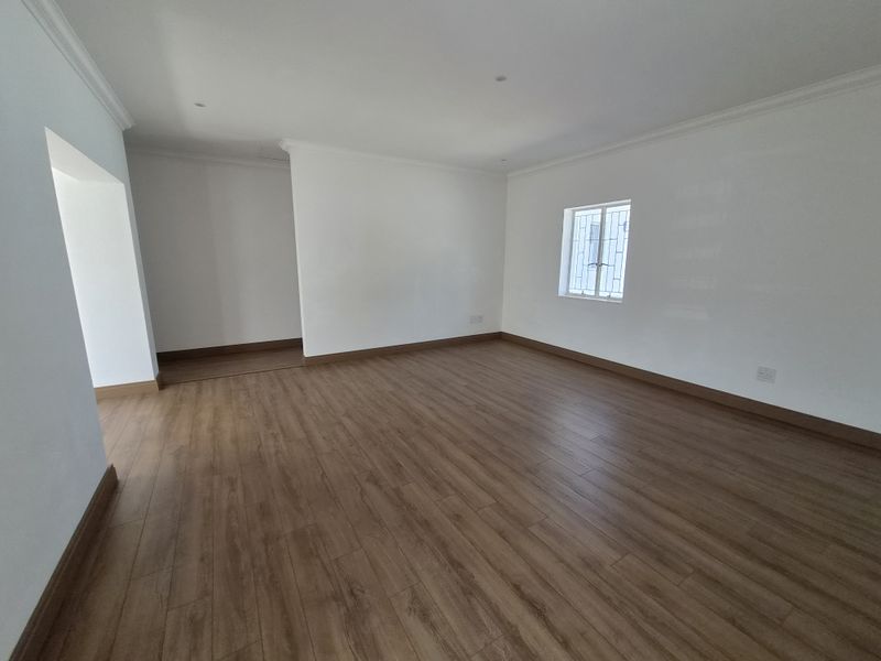 A bright, vacant living area featuring modern laminate flooring, clean white walls, coving, and a window fitted with security bars.