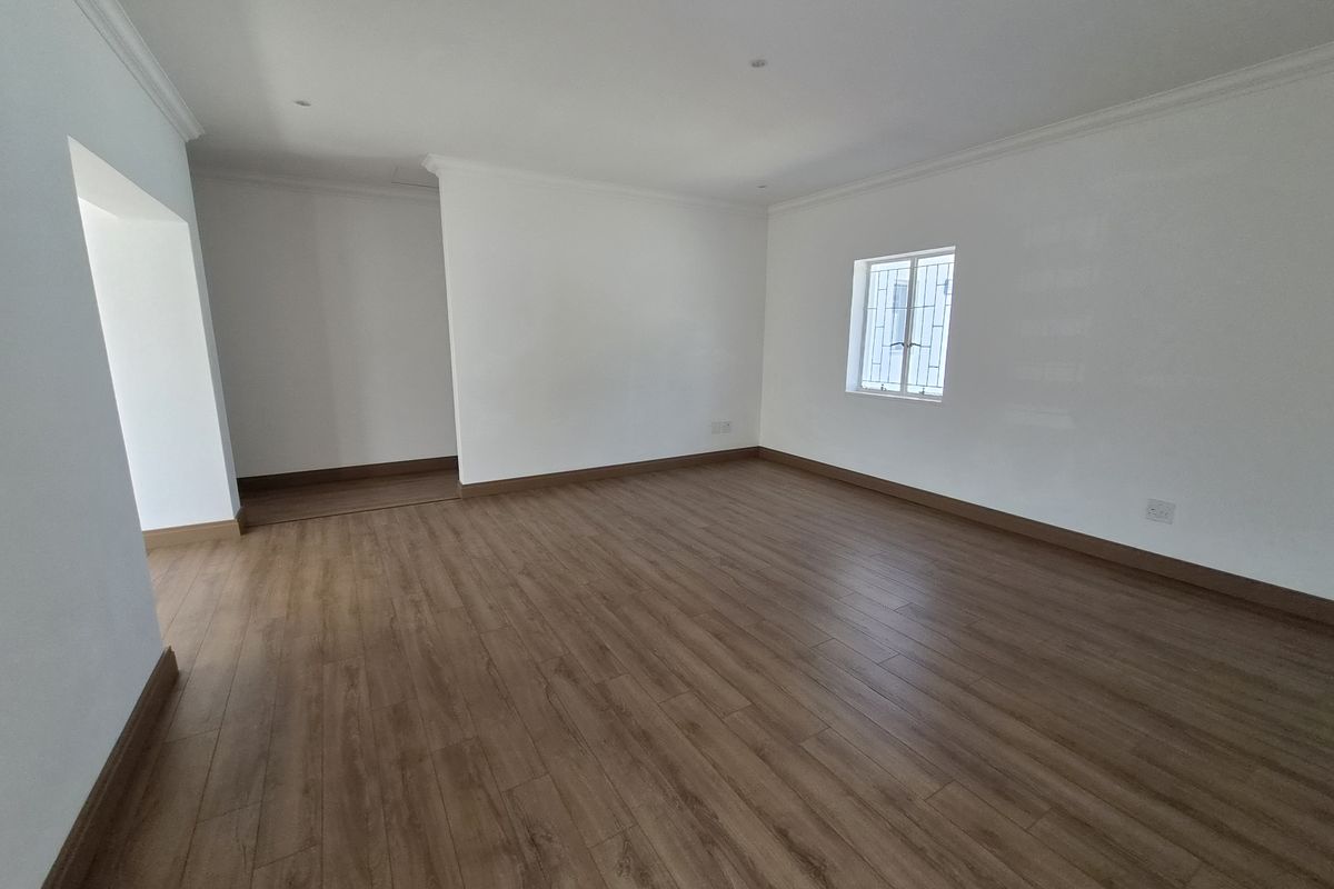 A bright, vacant living area featuring modern laminate flooring, clean white walls, coving, and a window fitted with security bars.
