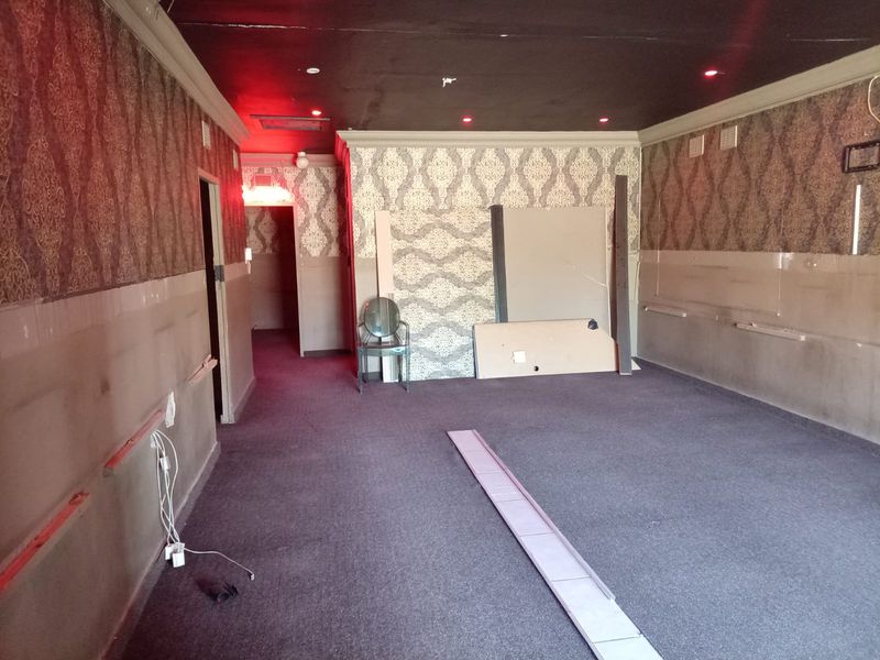 An empty commercial space featuring patterned wallpaper, dark carpeting, and recessed ceiling lighting, currently requiring cosmetic updates and repairs to wall finishes.