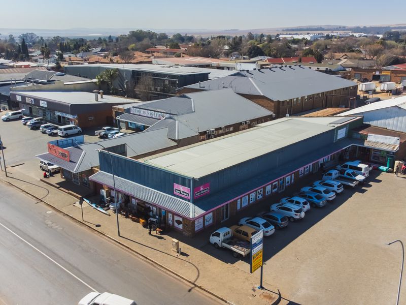 Aerial view of a commercial retail complex featuring multiple storefronts, metal roofing, and an extensive asphalt parking area.