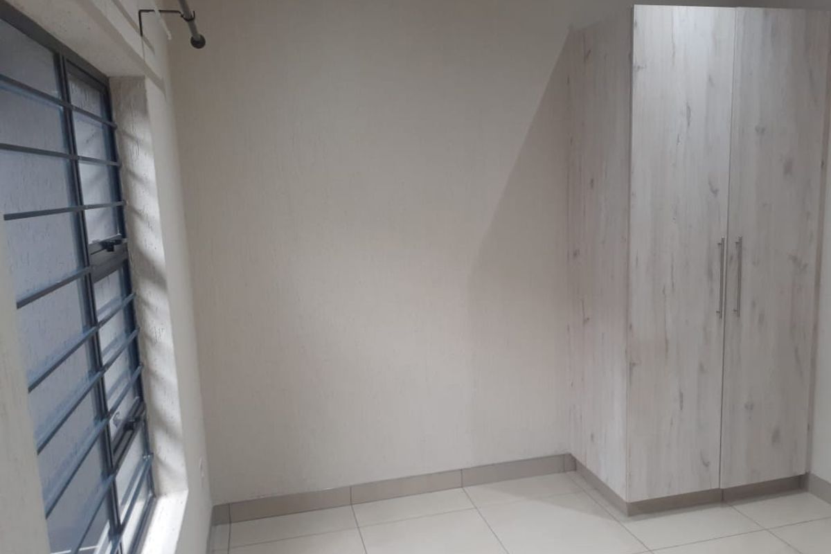 A clean, empty bedroom featuring neutral tiled flooring, a modern built-in wardrobe, and a window fitted with security bars.