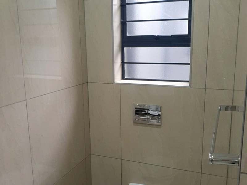 A modern, clean bathroom featuring a wall-mounted toilet with a concealed cistern, floor-to-ceiling tiling, and a window with security bars.