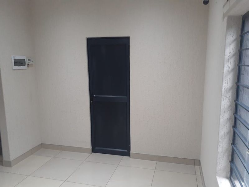 A clean, empty office space featuring neutral tiled flooring, a secure entrance door, and security bars on the window.