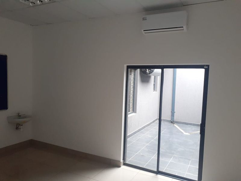 A clean, empty office space featuring tiled flooring, a wall-mounted air conditioning unit, a small basin, and a sliding glass door leading to a courtyard.