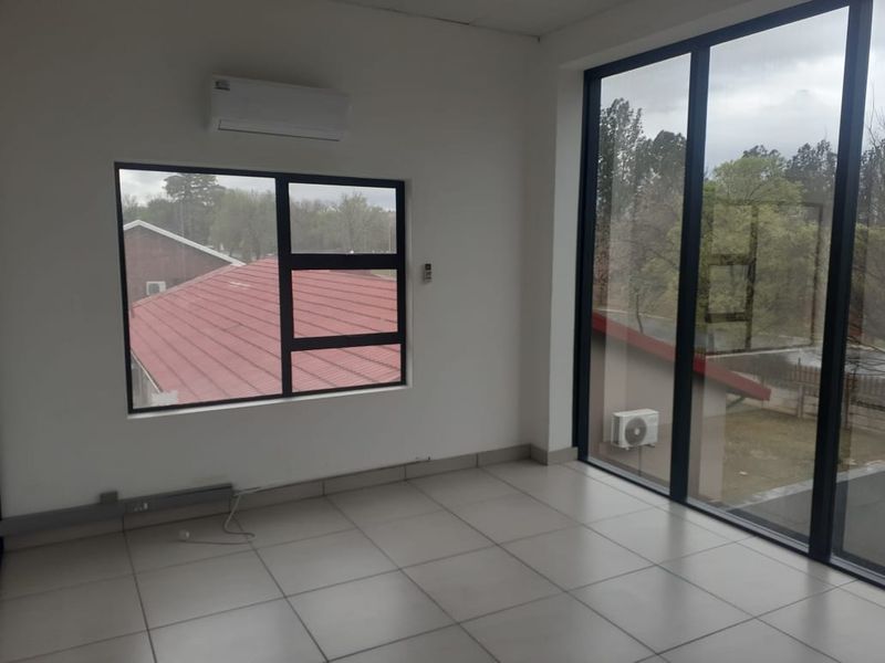 A bright, empty office space featuring large aluminium-framed windows, tiled flooring, and a wall-mounted air conditioning unit.